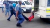 a man with his hands tethered to the back of a police vehicle being dragged behind as police hold his legs up and the vehicle apparently drives off, east of Johannesburg