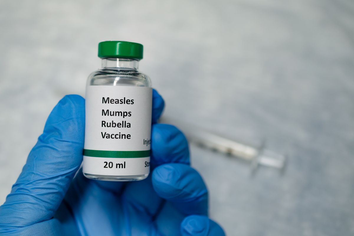 A hand wearing a surgical glove holds a vaccine vial that says “Measles Mumps Rubella Vaccine.” A syringe is visible in the background.