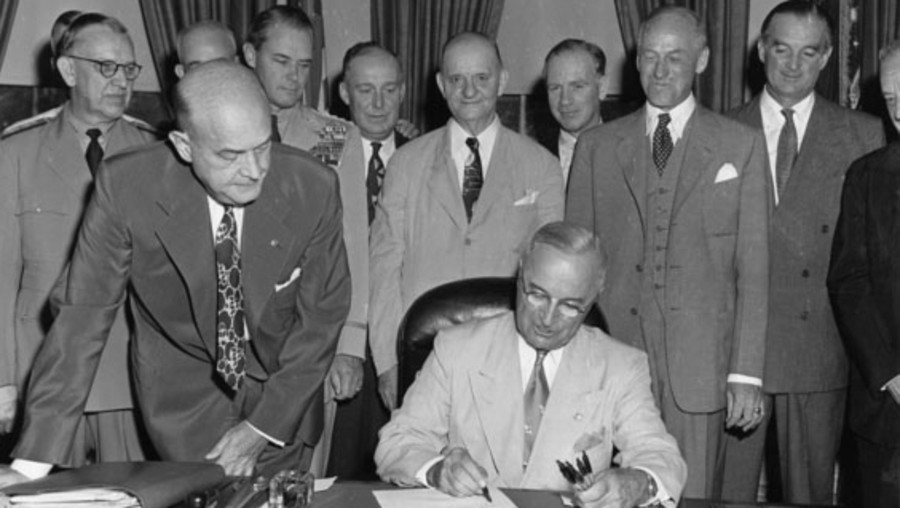 President Harry S. Truman signs a document at his desk in the Oval Office, as men stand around him and watch.