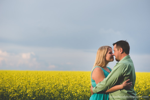 Pictured: a couple with noses touching and loving expressions on their faces, embracing in front of a bright yellow canola field and cloudy sky.