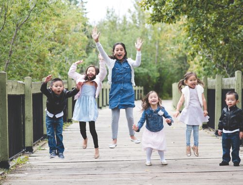 6 children at various stages of jumping in the air on a wooden bridge with green trees surrounding them