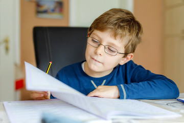 Portrait of cute school kid boy wearing glasses at home making homework. Little concentrated child writing with colorful pencils, indoors. Elementary school and education