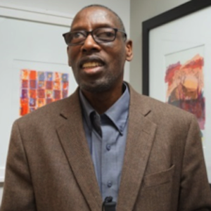 A photograph of Chester Finn. He is a black man with short, high salt-and-pepper hair. He is wearing glasses, a grey collared shirt and a course-woven brown suite jacket. Tow colorful orange paintings are in the corner of the room behind him.