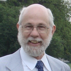 A portrait of Jack Derryberry. He has white hair and beard. He is wearing a pale blue suite and standing against a large green bush.