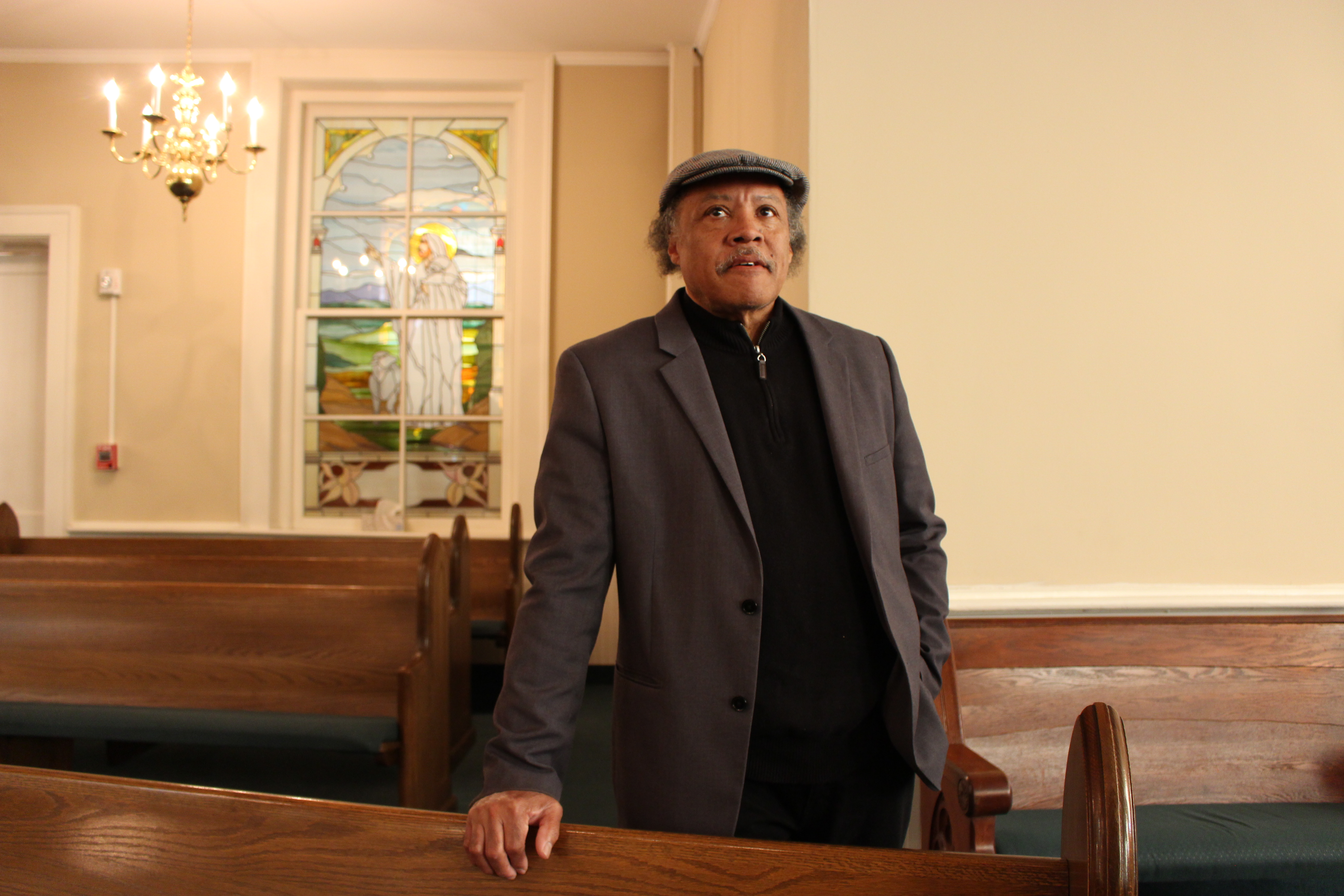 Elder Roy Harris stands in his church, Mt Zion Missionary Baptist Church for a portrait. He rests his right hand on a pew and looks beyond the camera towards the front of the worship hall. He is wearing a grey suit, black sweater, and a grey flat cap hat.
