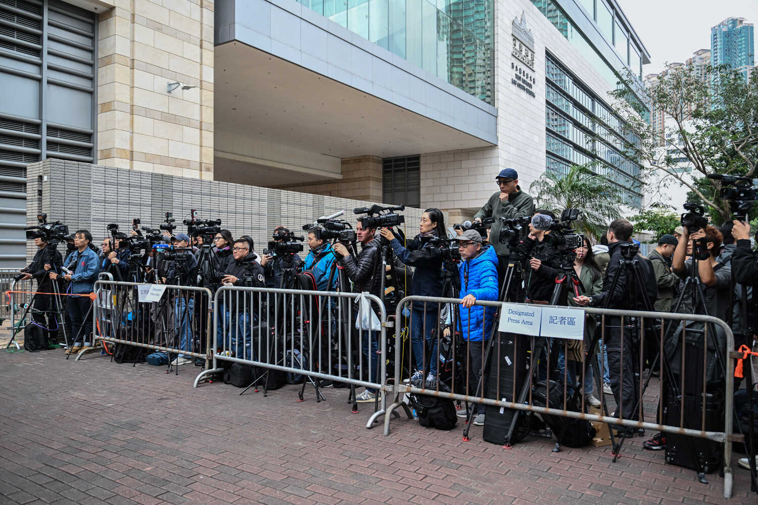 Journalists crowd together, their cameras pointed in the same direction, behind a fence set up outside a building.