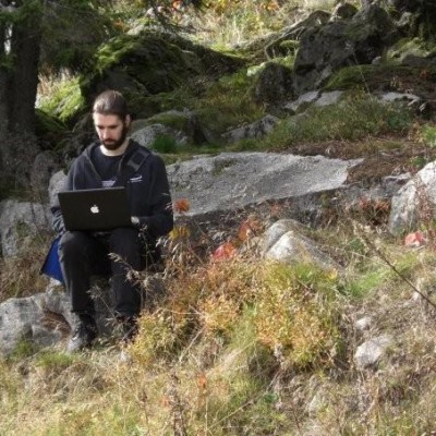 Photo of Ingmar sitting on a rock in a pine forest with eyes focused on his grey mac laptop